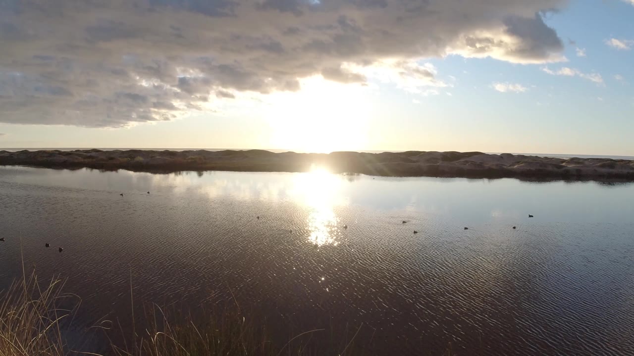 A wide view of the lake in Pismo Beach shows the sun reflected near center, a low dark shoreline, pale blue sky, large clouds, and no visible people.
