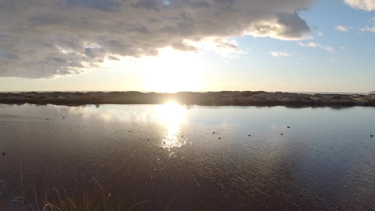 Sunlight flares across the left-center water on a Pismo Beach lake, with cloud reflections, a shadowed shoreline ridge, small dark specks on the water, and no visible people.