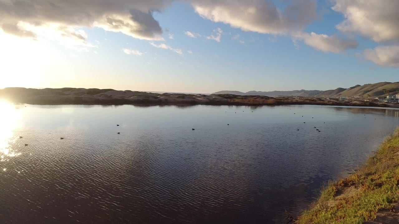 Pismo Beach overlook with calm rippled water across the frame, low hills on the horizon, warm sunlight from the left, and no visible people at 0:01.