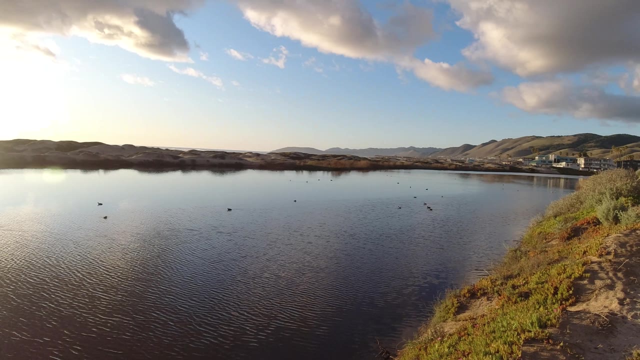 Wide Pismo Beach frame showing rippled water, distant brown hills, heavy clouds, a bright left-side sun reflection, and scrub-covered shoreline with no visible people.
