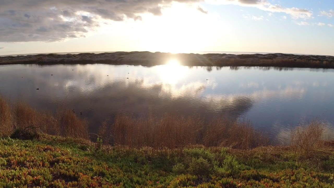 At Pismo Beach, California, bright sunrise glare sits near the distant ridge and mirrors in the lake, with darker cloud shapes above and no visible people.