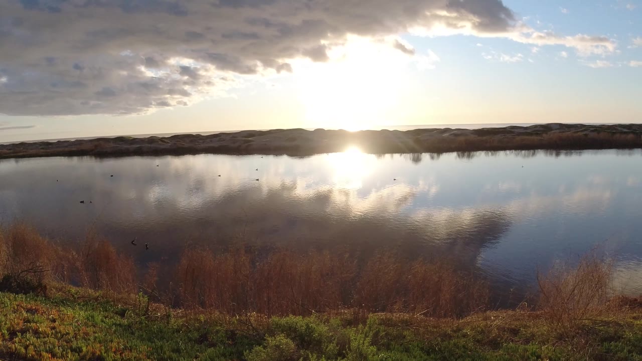 At Pismo Beach, California, cloud cover spreads across the upper left while the sun and clouds reflect on the lake, with brown wetland plants below and no visible people.