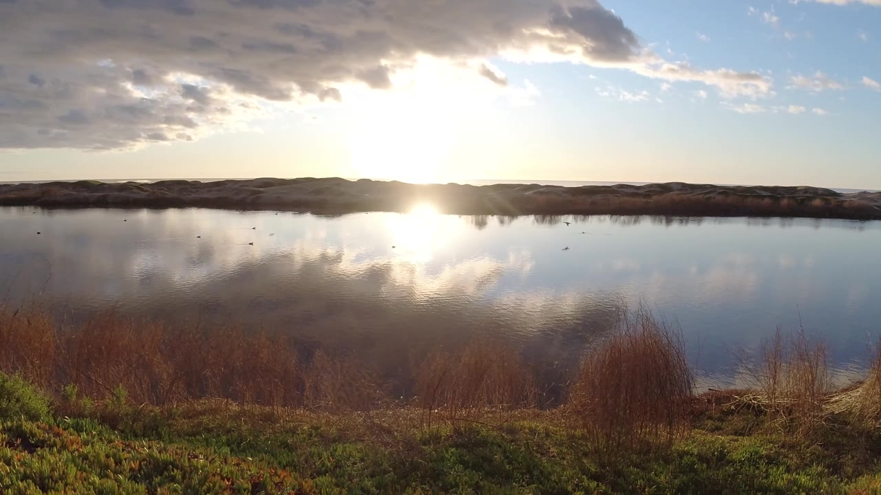 Pismo Beach lake-bank frame with sun glare on the water, a dark ridge on the horizon, thicker foreground grasses at right, and no visible people.