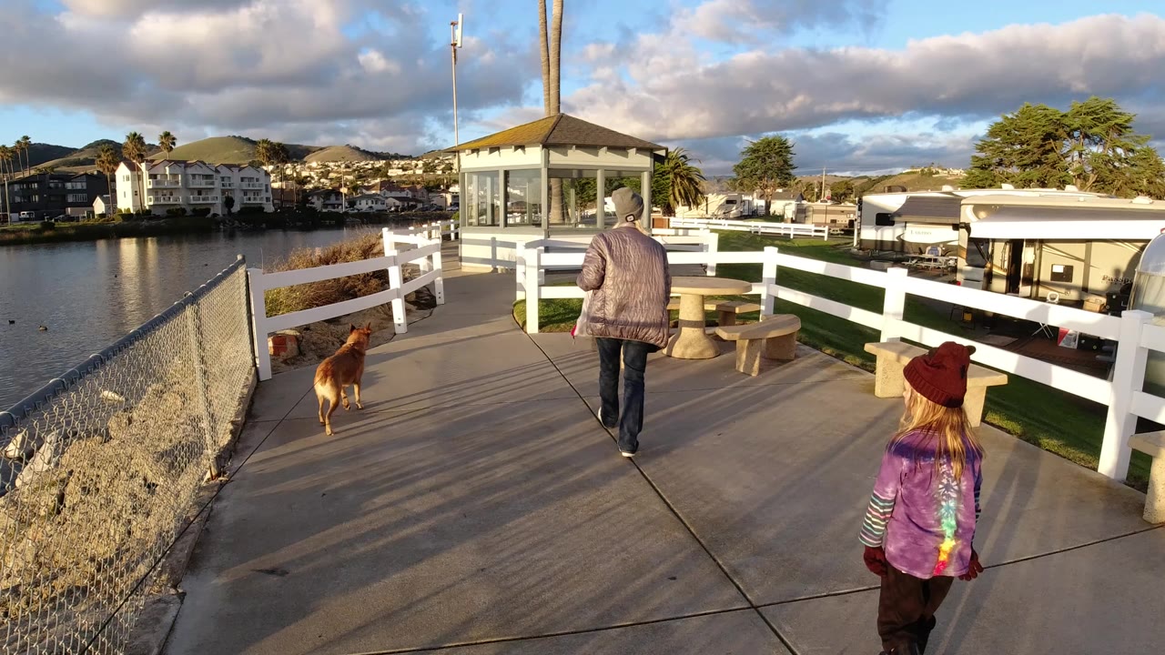A woman, child, and brown dog walk away from the camera on a Pismo Beach waterfront boardwalk with white fencing, benches, water, houses, hills, and warm low sunlight.