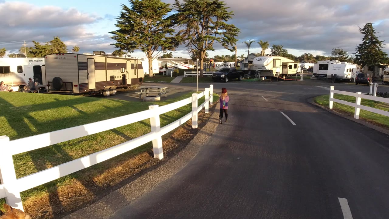 A child runs ahead on a paved lane in Pismo Beach, California, with a white fence in the foreground and RVs, trailers, trees, and warm sunlight behind.