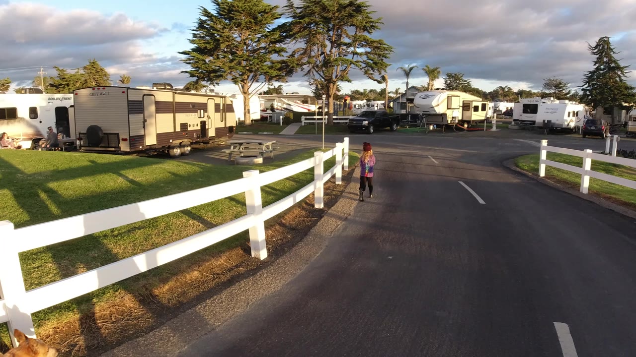 The child continues along the Pismo Beach RV park road while a white fence crosses the foreground and sunlit trailers and tall trees fill the background.