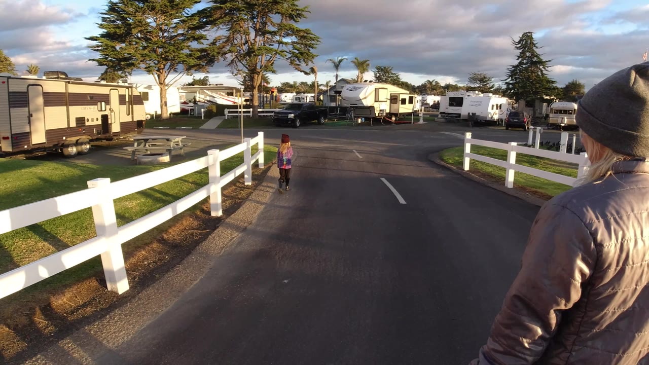 A blond adult in a gray beanie appears at the right edge as the child runs ahead on the Pismo Beach road, with RVs, a white fence, and late-day shadows visible.