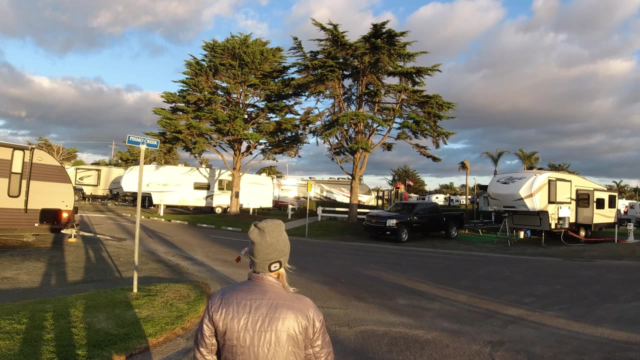 At 0:00 in Pismo Beach, a person in a gray beanie and reflective jacket walks away from the camera on a paved street lined with RVs, trees, and long shadows.