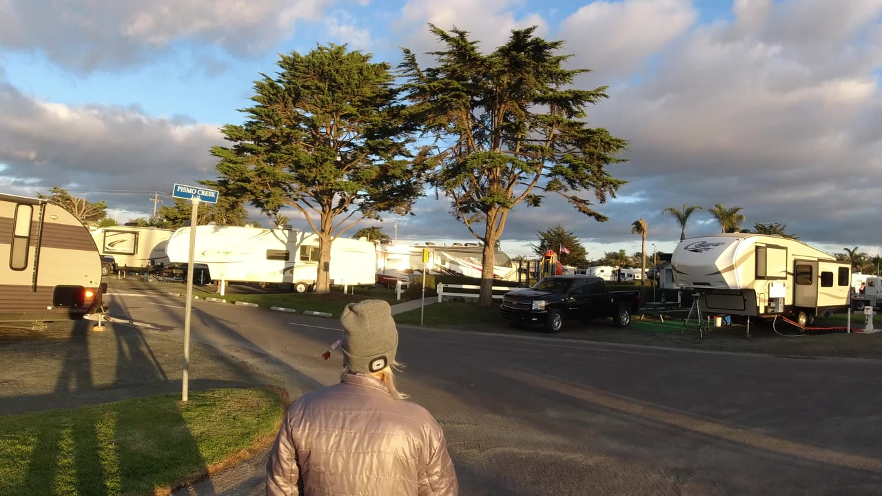 At 0:01 in Pismo Beach, the walker remains centered from behind while RVs, a black pickup truck, curbside grass, trees, and low sun fill the street scene.