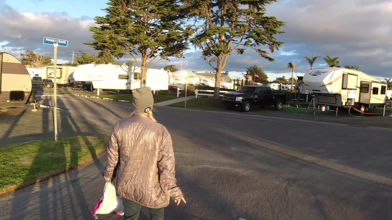 At 0:02 in Pismo Beach, the person carrying a white bag moves farther along the road, with a cross street, parked RVs, palm trees, and a black pickup visible ahead.