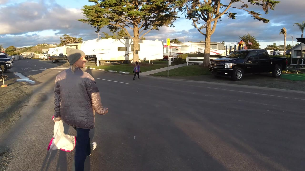 In Pismo Beach, California, a person in a knit cap and shiny coat steps from the near side of the street holding a white plastic package, with RVs, parked vehicles, trees, and a child visible ahead.