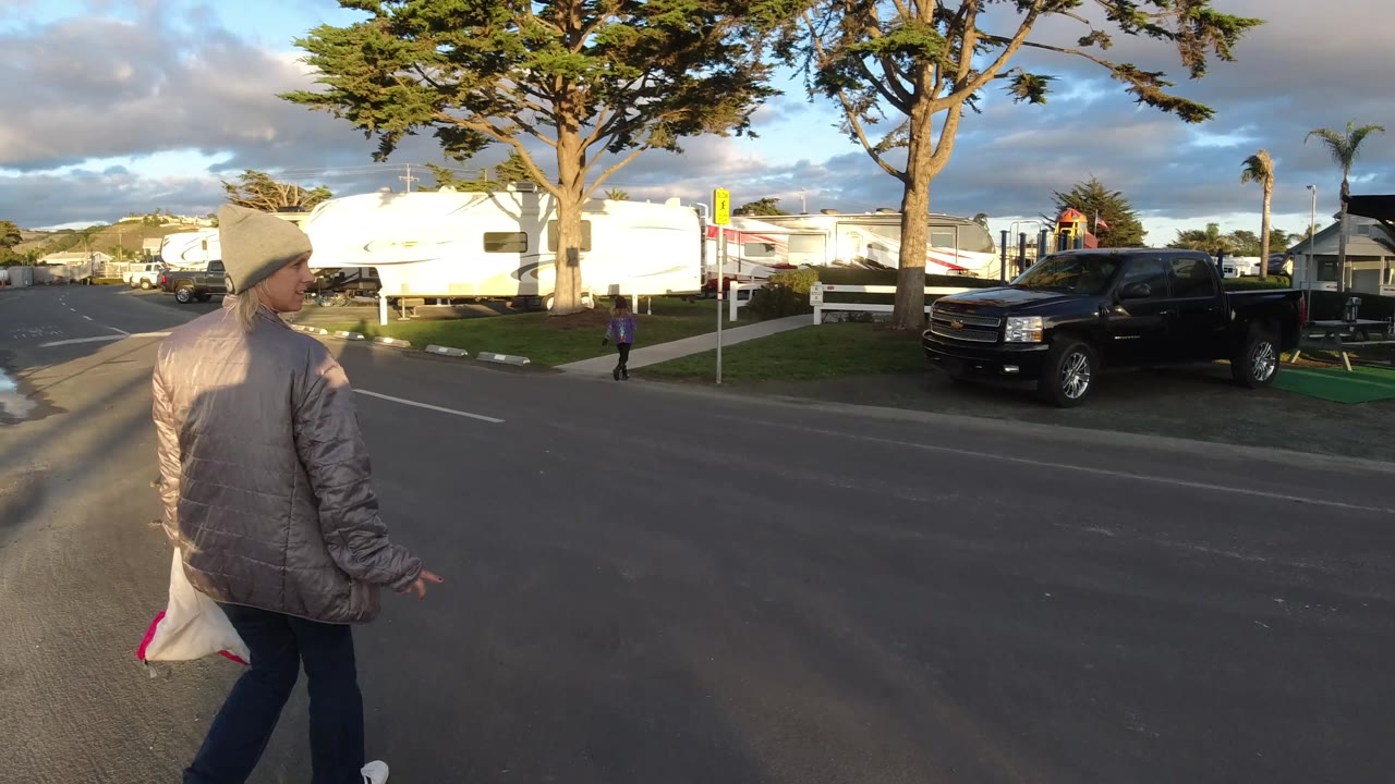 In Pismo Beach, California, the foreground person continues across the street in warm side light, with a black truck parked near the curb, white RVs behind a fence, trees overhead, and a child near the sidewalk.