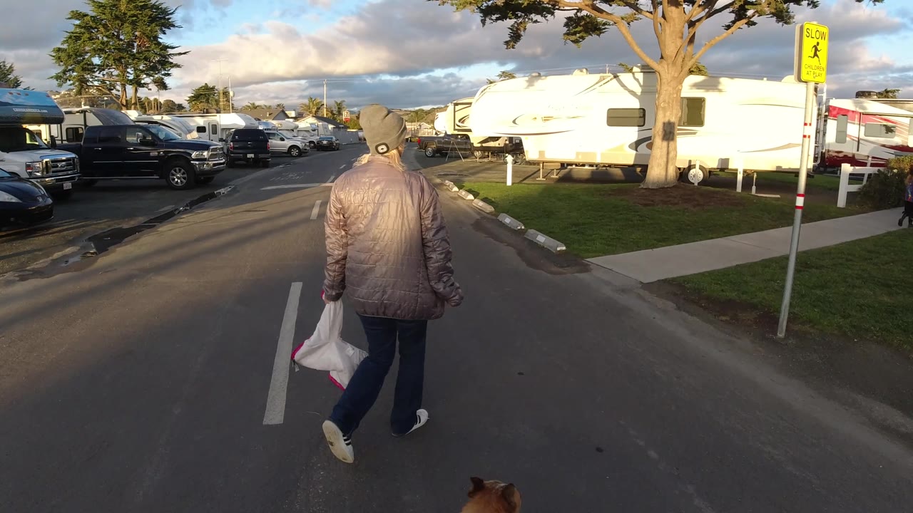At 0:02 in Pismo Beach, the man walks away near the right lane marking, with a yellow slow-children sign, RVs, parked vehicles, grass, a sidewalk, and a partly cloudy sky visible around him.