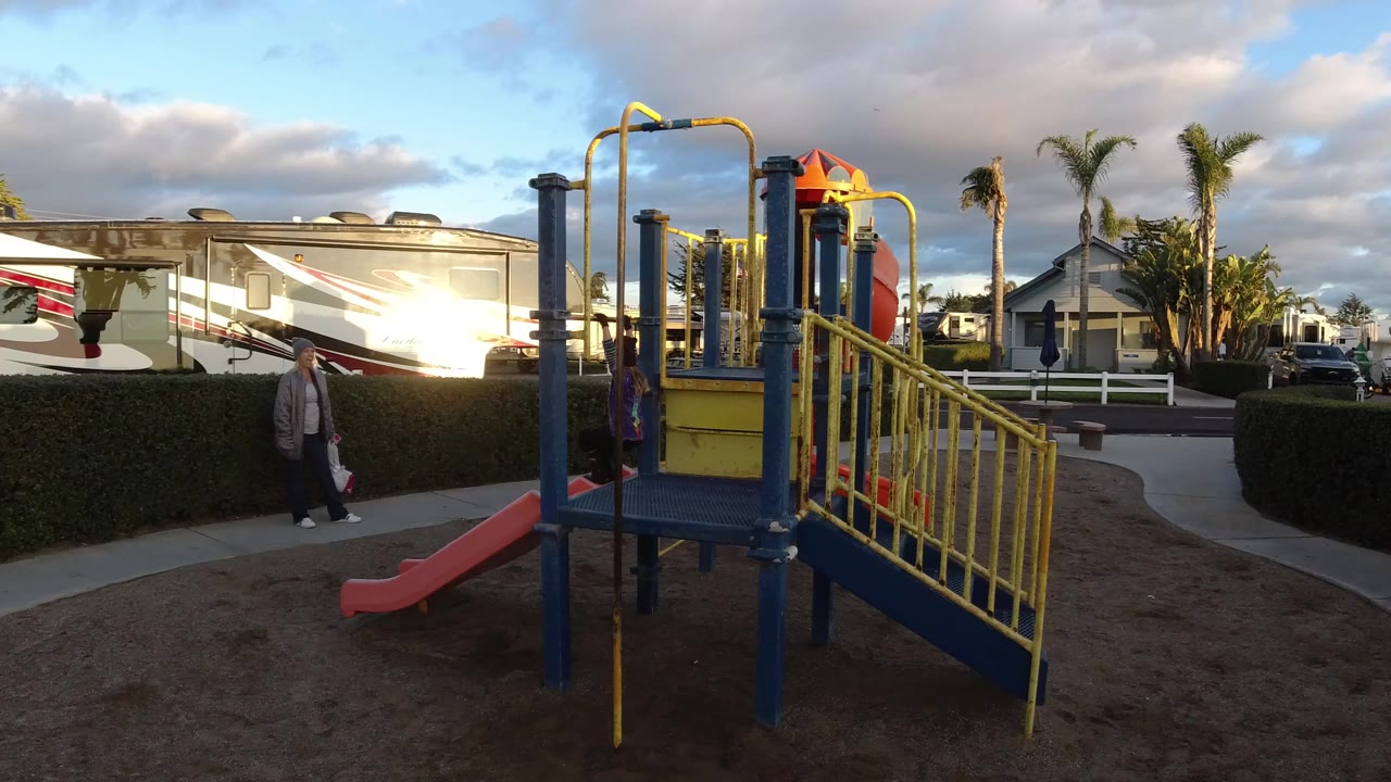 Wide view of a blue and yellow playground in Pismo Beach with an adult and child near the walkway, RVs and palm trees behind the hedge.