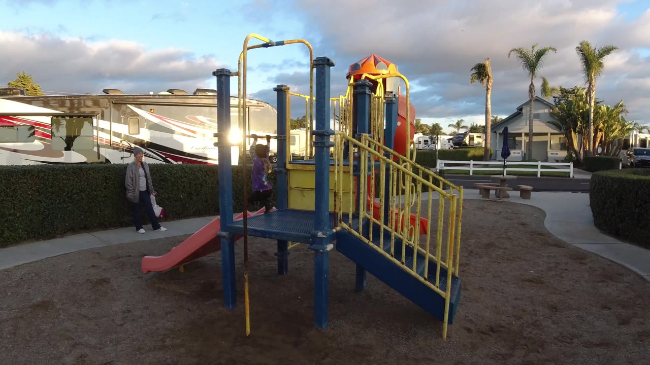 A wider Pismo Beach park view shows yellow playground stairs and a red slide in the foreground, with an adult and child near the hedge and RVs beyond.