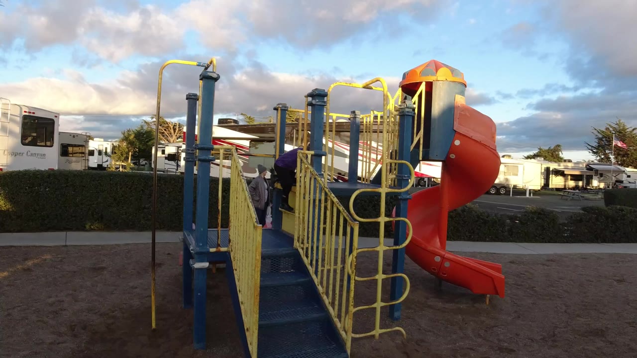 Wide view of a Pismo Beach playground with blue stairs, yellow railings, an orange spiral slide, RV trailers behind the hedge, and a child partly visible on the play structure.