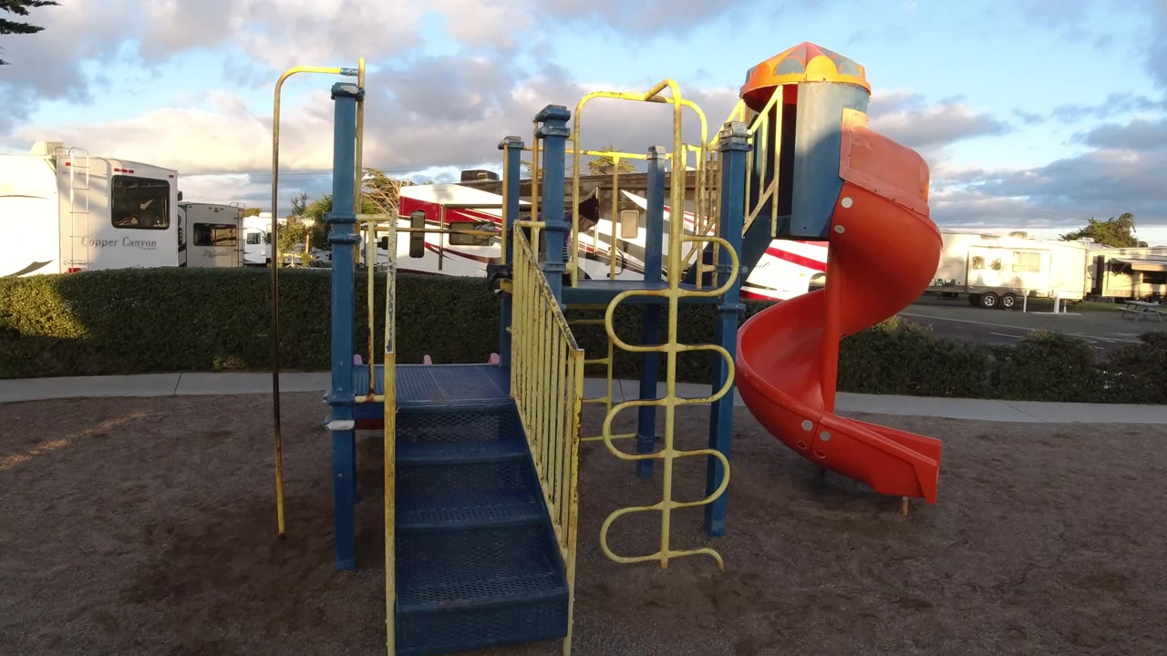 Wide Pismo Beach playground view with blue stairs, yellow railings, an orange slide, hedges, and RV trailers in the background, with no visible people in this frame.
