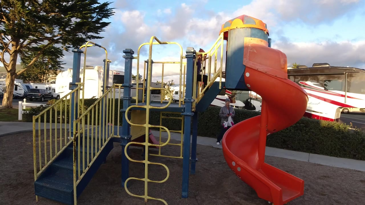 At 0:02 in Pismo Beach, the slide remains prominent while an adult appears near the sidewalk beside the hedge and a child is partly visible behind the play structure.
