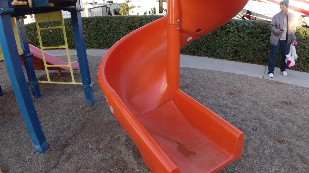 Orange slide and yellow ladder over sand with an adult in the background.
