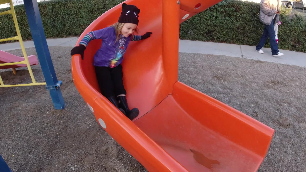 A child in a dark knit hat, purple shirt, gloves, and boots reaches the lower curve of the orange slide at the Pismo Beach playground.