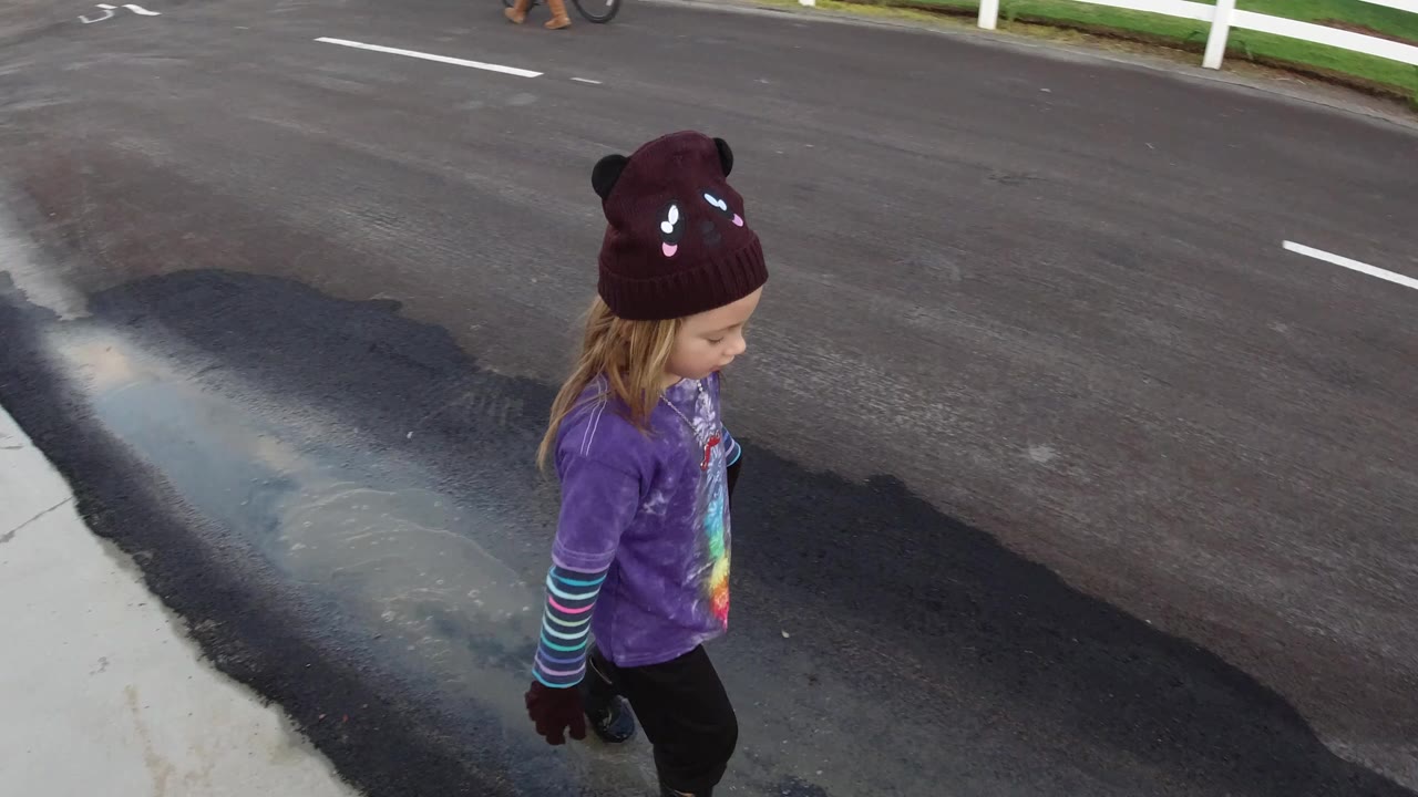 A child in a burgundy knit hat and purple shirt walks along a paved road in Pismo Beach, California, with a dark wet patch on the asphalt nearby.