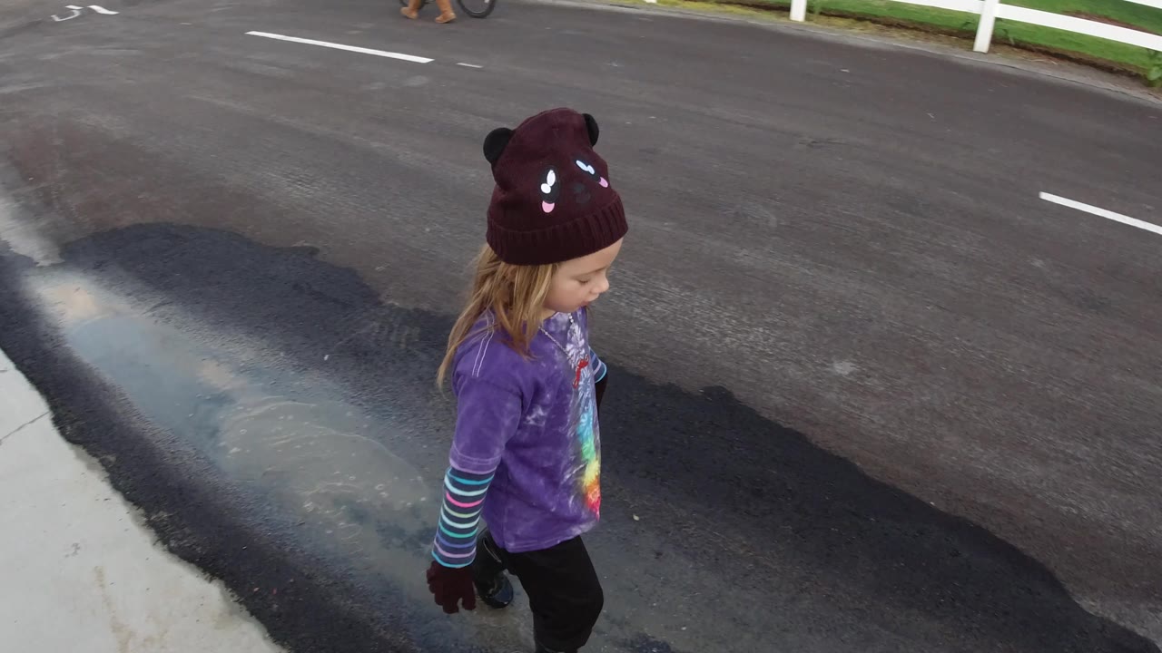 The child continues walking on the Pismo Beach road, seen from above with the wet asphalt patch and lane markings filling most of the frame.