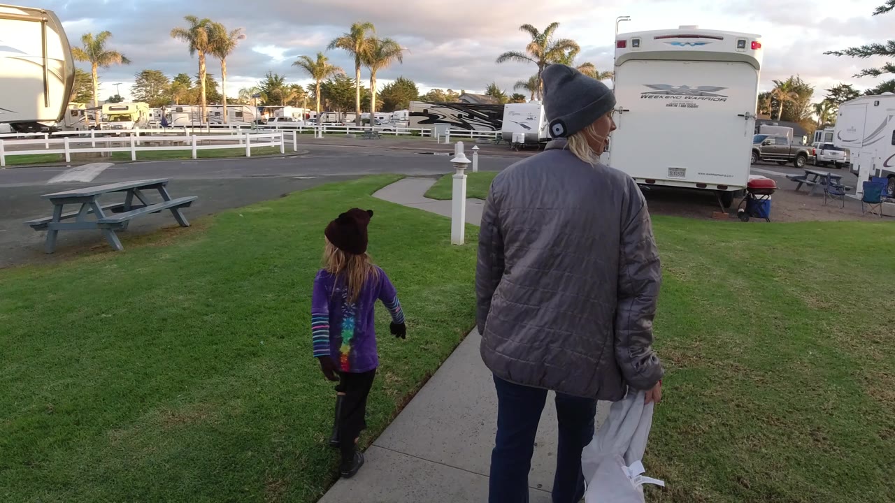 The child and woman continue forward on the Pismo Beach sidewalk, with a large white RV close on the right and rows of parked trailers beyond the white fence.