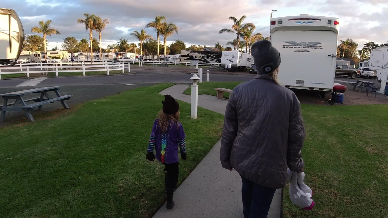 At 0:02, the woman and child are farther along the sidewalk in Pismo Beach, with the large RV rear filling the right side and open grass and fencing to the left.