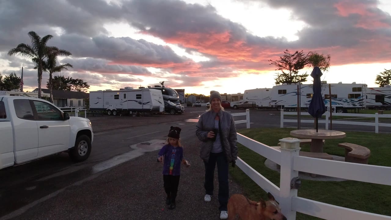 In Pismo Beach, the woman and child continue along the RV park road while the dog is partly visible at the bottom edge and a white pickup sits near the fence.