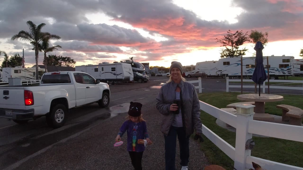 In Pismo Beach, a white pickup drives through the frame near the woman, child, and dog, with RVs, fencing, and a bright strip of sunset below heavy clouds.