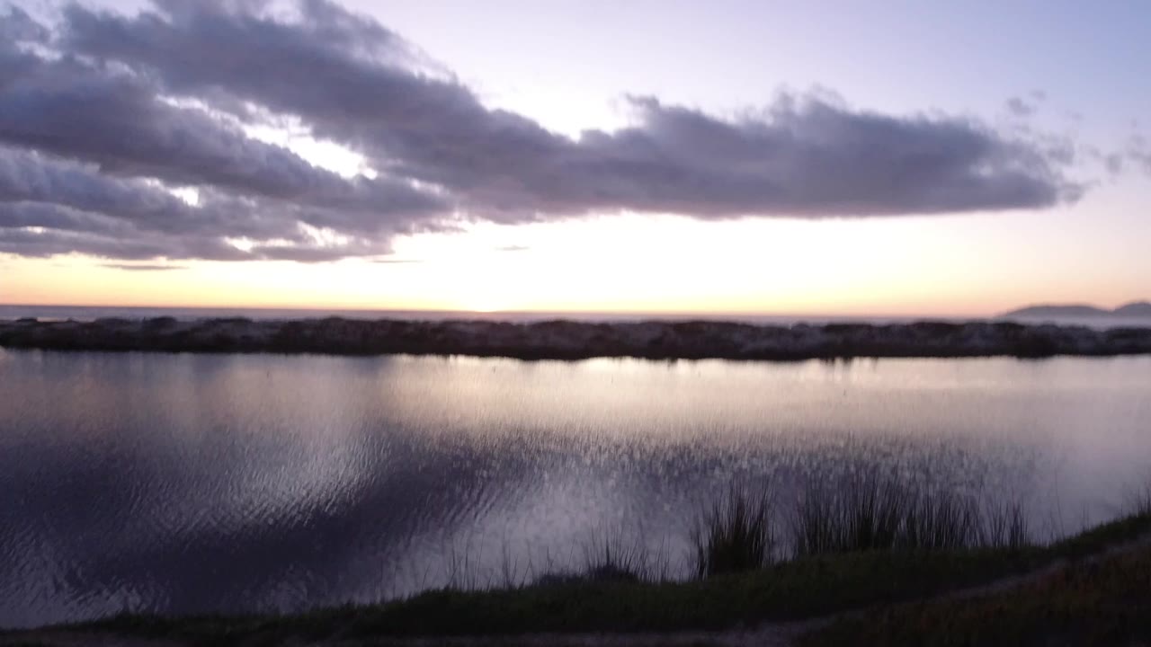 Pismo Beach, California water edge at late afternoon with rippled reflections, a low rock line, distant hills to the right, blue-gray clouds, and no visible people.