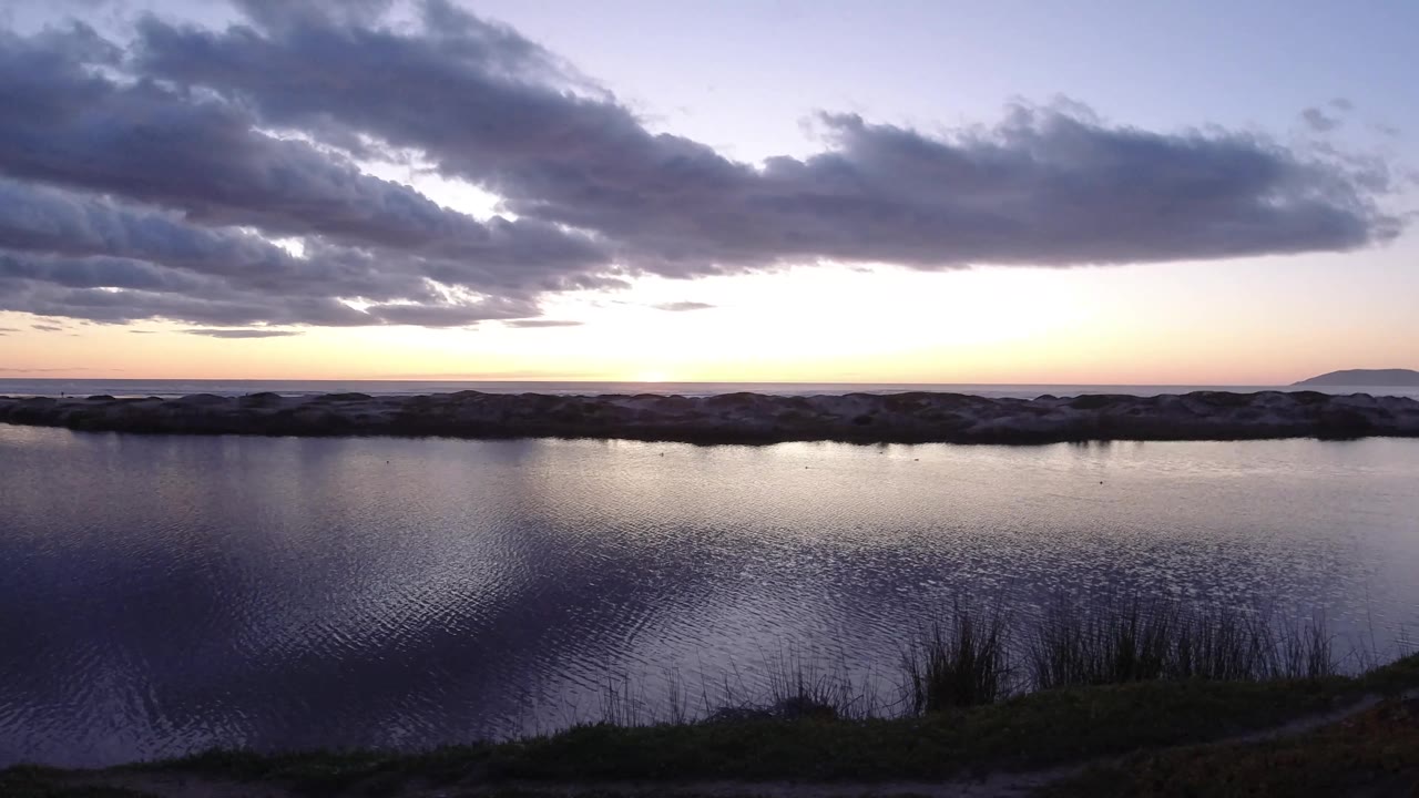 Pismo Beach, California sea-edge scene with textured ripples, reeds near the lower frame, a dark cloud band overhead, and no visible people.