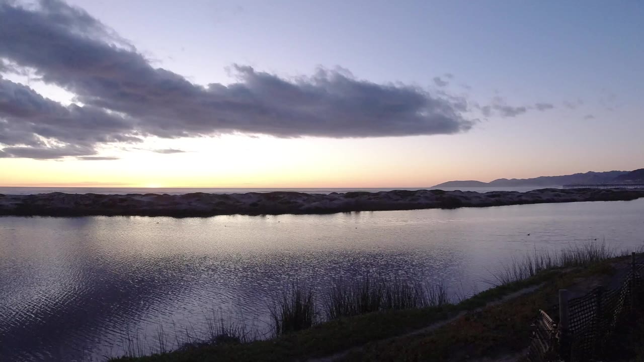 Pismo Beach sunset view across rippled water, with reeds near the lower edge, a dark rock line, distant mountains to the right, and no visible people.