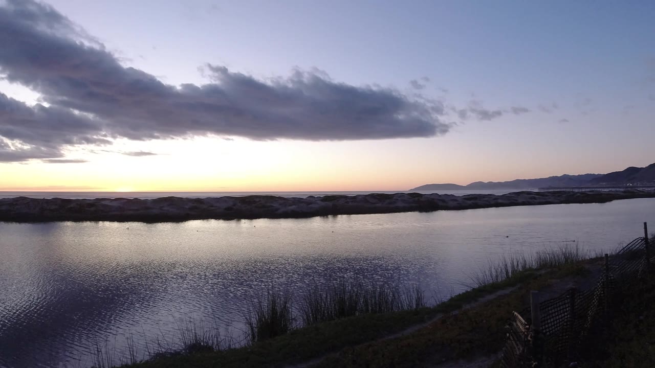 Pismo Beach sunset frame with a darker cloud stretched across the upper sky, calm water below, reeds at the bottom edge, and no visible people.