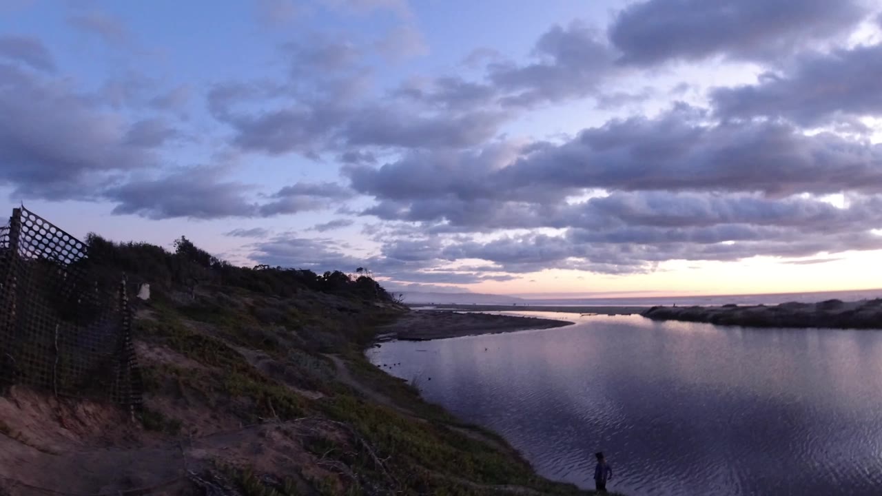 Pismo Beach, California shoreline at dawn with calm reflective water, dark dunes, a lattice fence on the right, heavy clouds overhead, and a small silhouetted person near the lower edge.