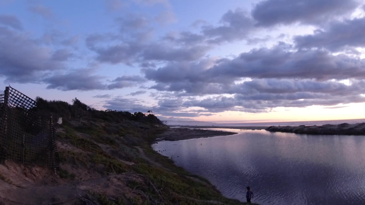 Pismo Beach shoreline view with the water centered, purple-blue clouds across the sky, a dark fence and dunes at the right edge, and a distant person low in the frame.