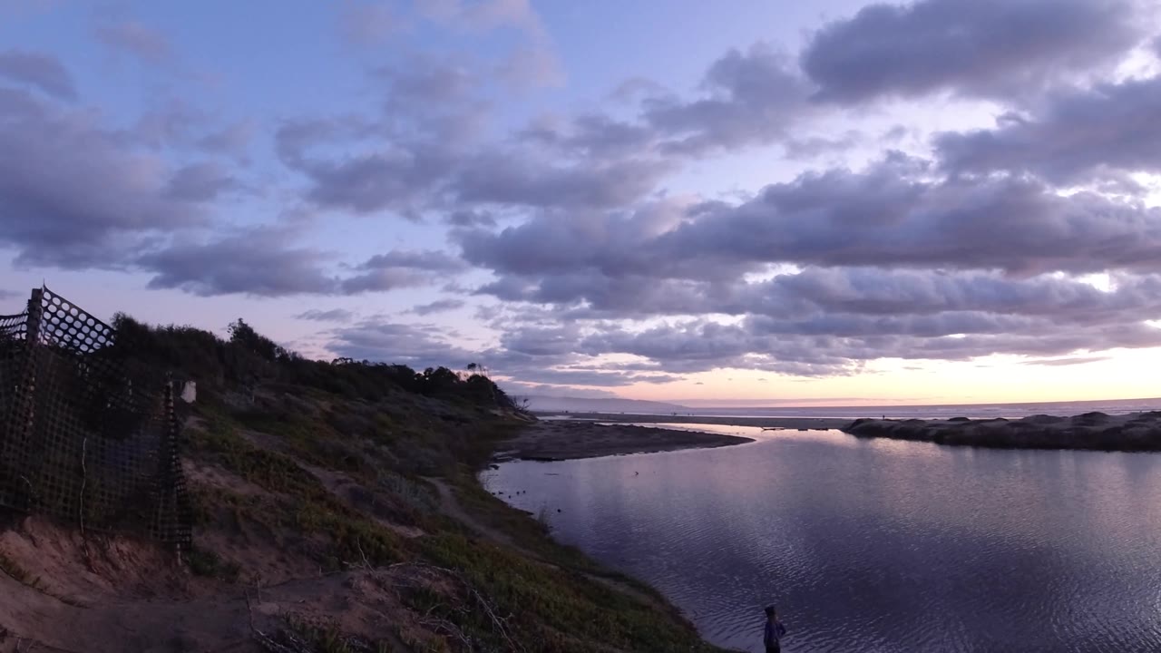 Pismo Beach coastal water under heavy dawn clouds, with a pale horizon band, dark dunes and brush, a fence on the left side of the view, and a small person near the lower shoreline.
