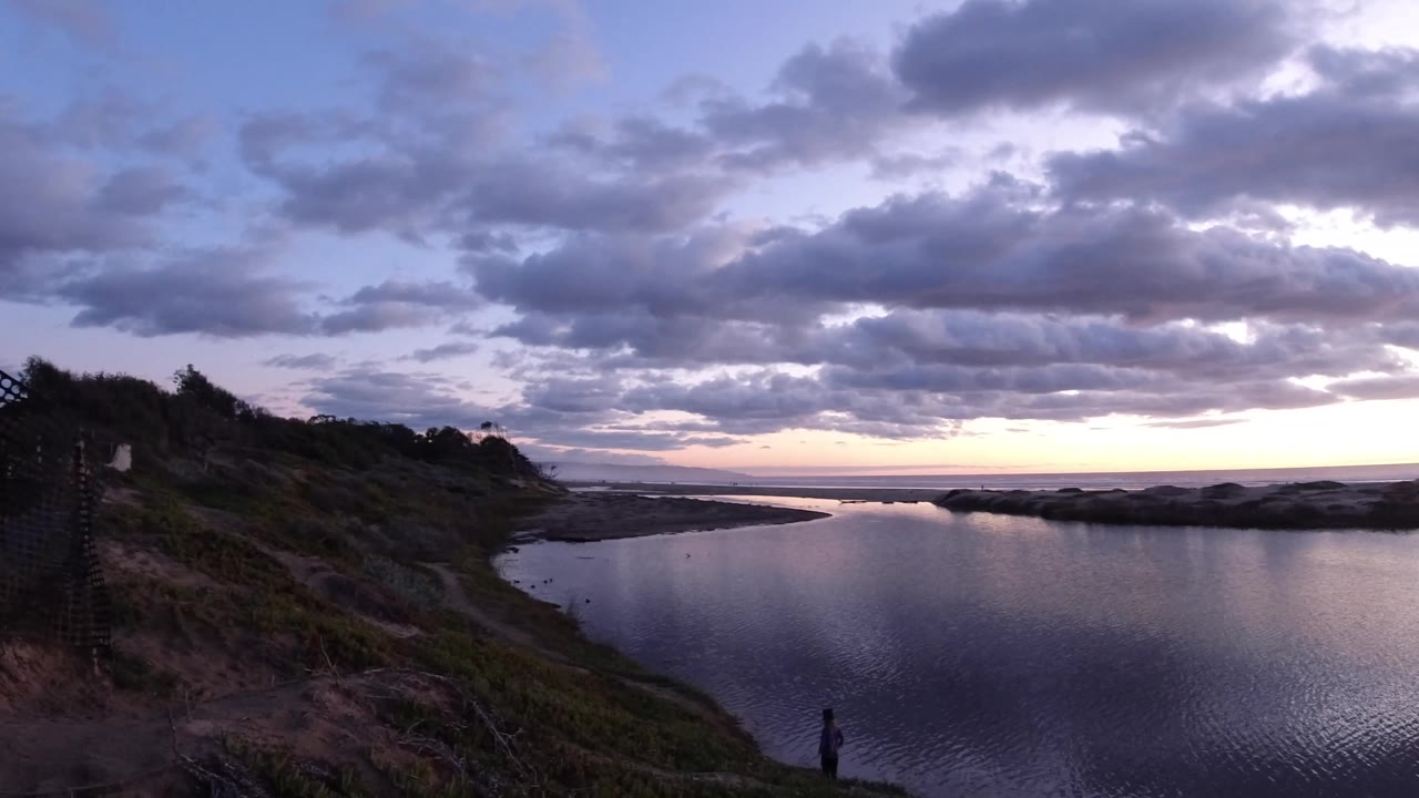 Wide shoreline water scene at dawn with clouds, dunes, and a distant walker.