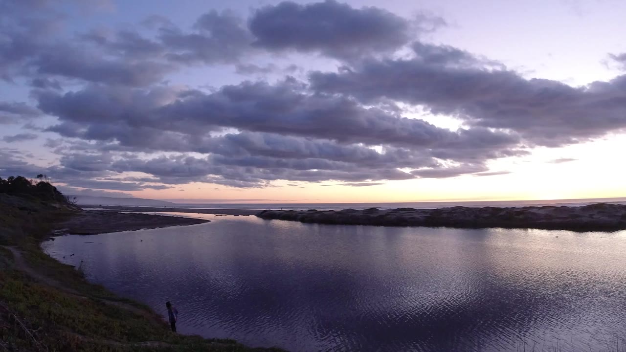 Wide Pismo Beach sunset view with dark rippled water in the foreground, a low shoreline across the center, and purple gray clouds above a thin orange horizon.