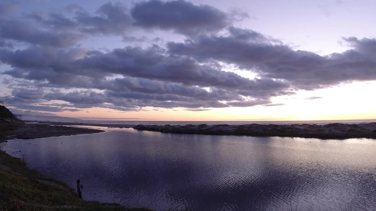 Wide Pismo Beach dusk view with the water foreground pushed dark, rocks or dunes across the middle, and a bright pale band below dense blue gray clouds.