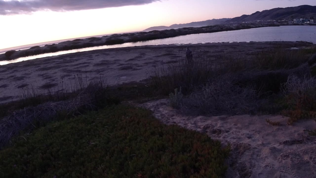 Pismo Beach appears in a wide elevated view with shadowed scrub in front, muted sand flats, a band of water, distant hills, and a bright sunset edge at left.