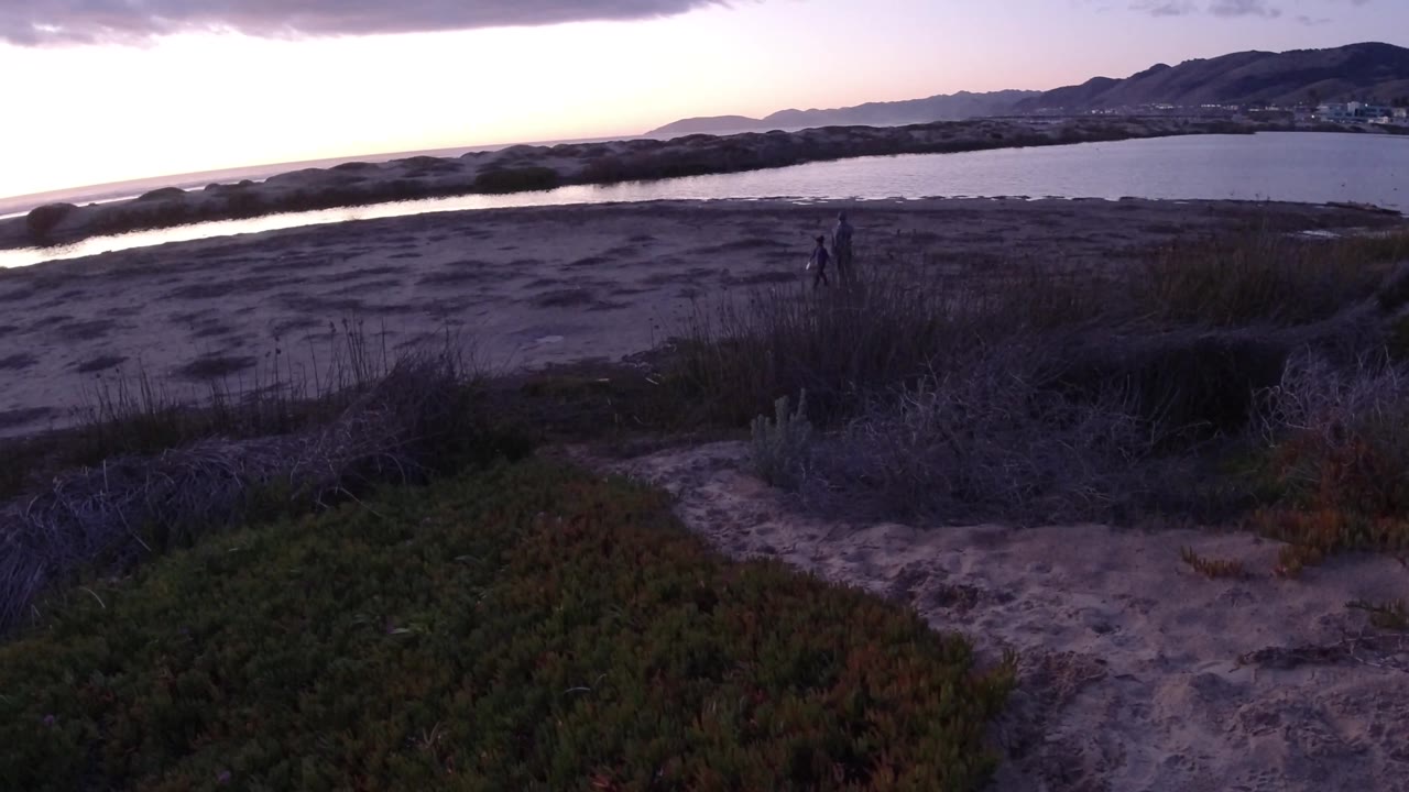A Pismo Beach sunset view shows low brush and sand in the foreground, water behind the beach, hills on the horizon, and two tiny distant human-like silhouettes near the midground.