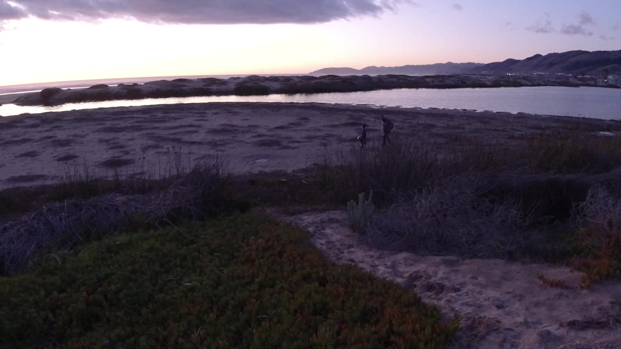 Dusk beach view with two small distant silhouettes on sand near water and hills beyond.