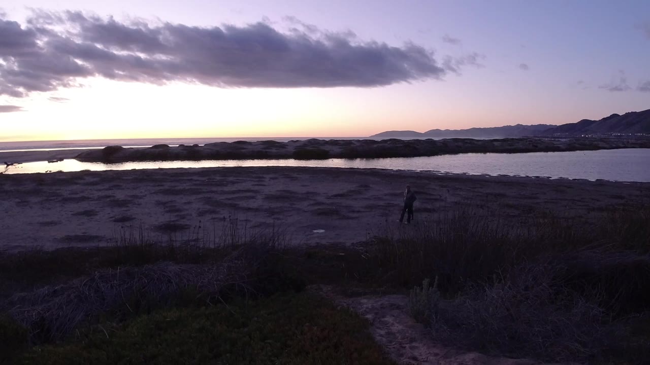 Pismo Beach dusk frame with the adult and child slightly right of center on the shoreline, water behind them and a bright band of sky near the horizon.