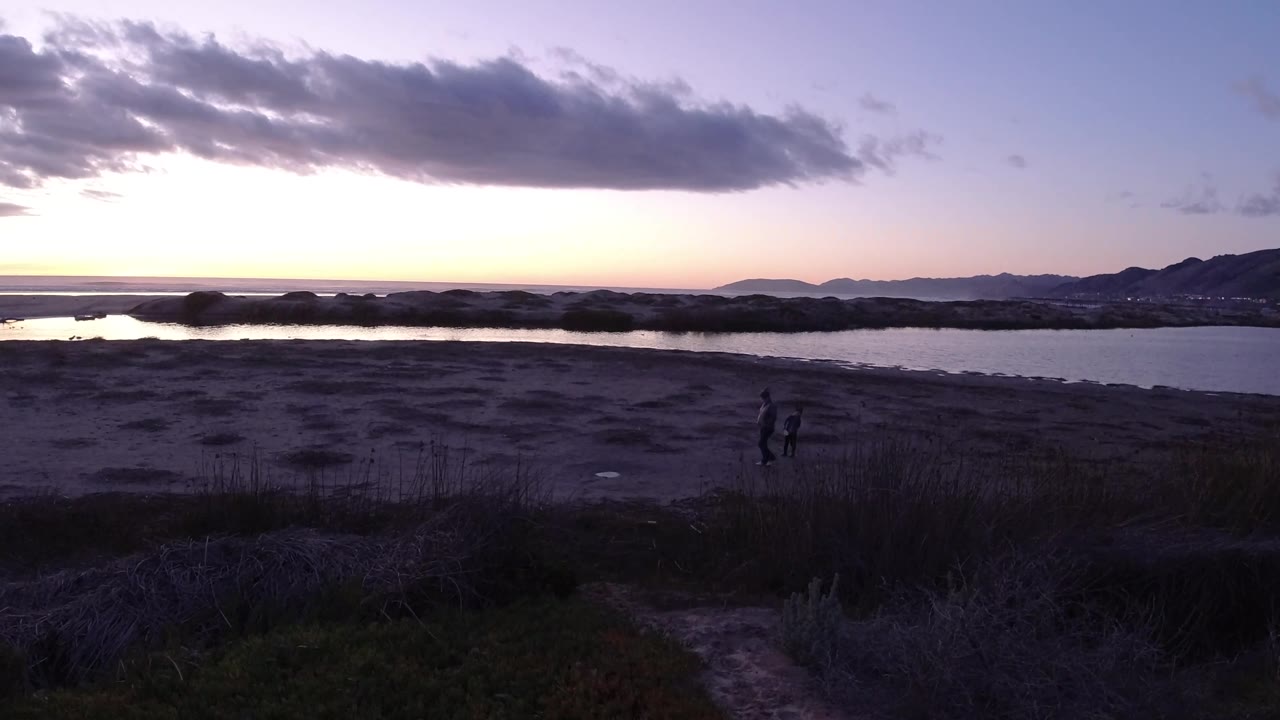 Wide Pismo Beach view with two small people near the right edge, dark beach foreground, reflective river water, and hills under purple-blue dusk sky.