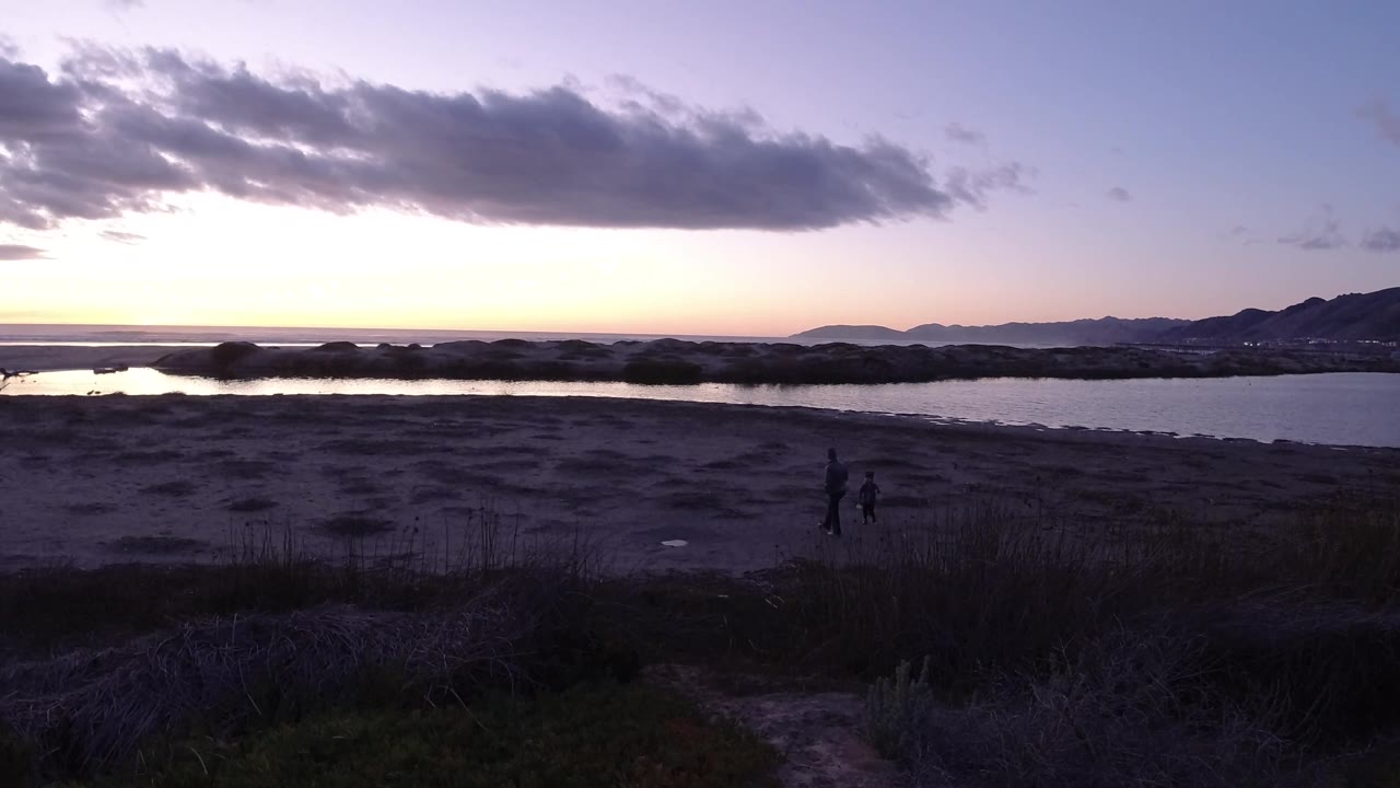 Two small figures walk along a shadowed sandy bank at dusk.