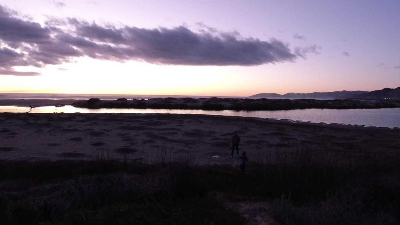 Wide dusk view in Pismo Beach, California, with dark dunes in front, calm water beyond, low hills on the horizon, heavy clouds overhead, and small people visible near the lower foreground.