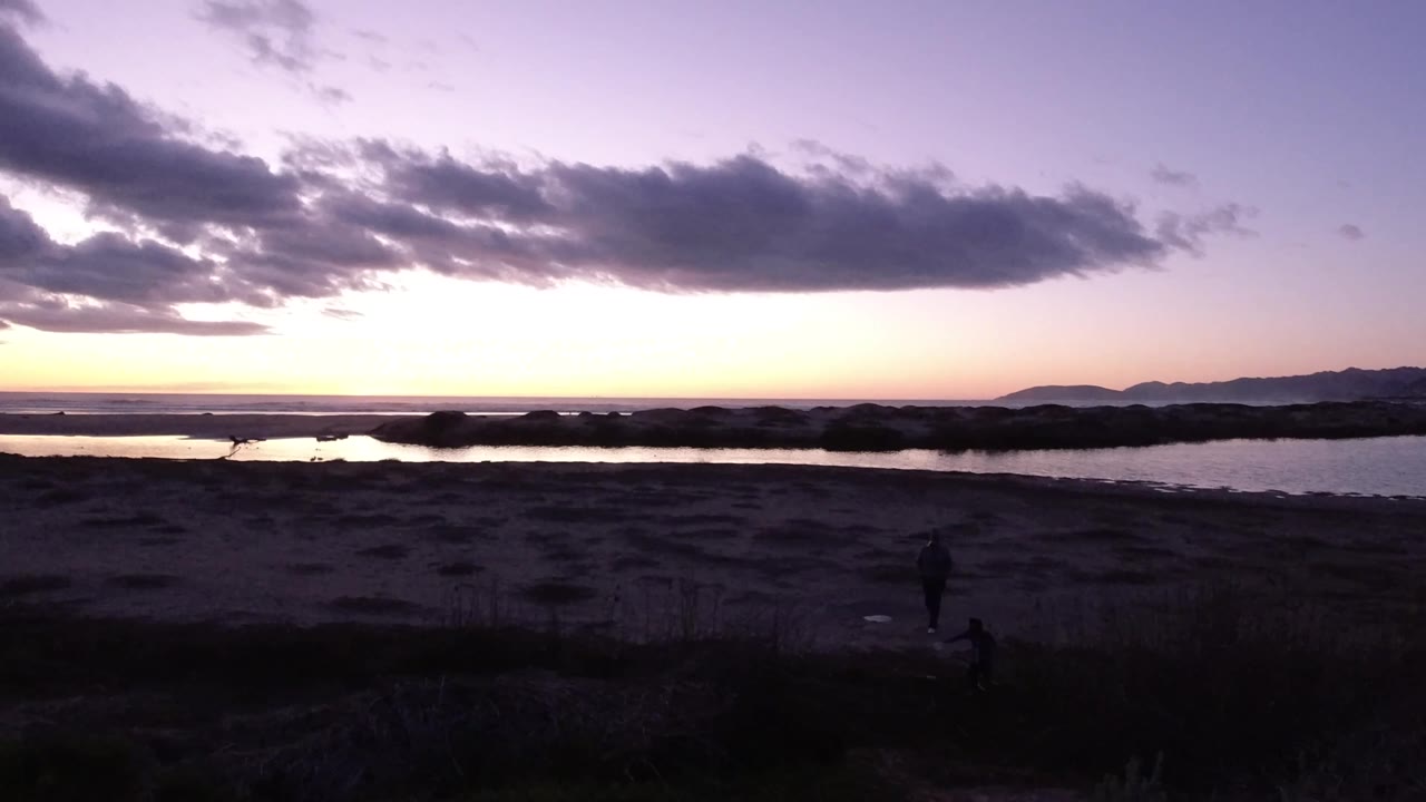 Wide Pismo Beach shoreline at dusk, showing calm water under a peach horizon, a large dark cloud overhead, shadowed sand and brush, distant hills, and a small person near the lower right.