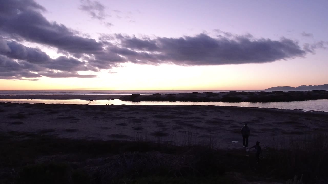 Low-light beach and water at dusk with clouds, hills, and a small person at the lower right.