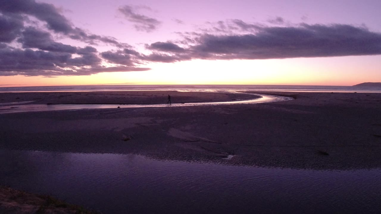 Wide view of Pismo Beach at sunset with rippled foreground water, dark sand flats, a winding tidal channel, a distant headland, and a small person visible near the channel.